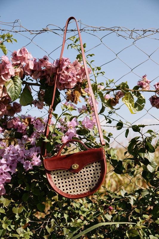 Brown woven handbag with a pink strap hanging on a wire fence with pink flowers and green leaves.