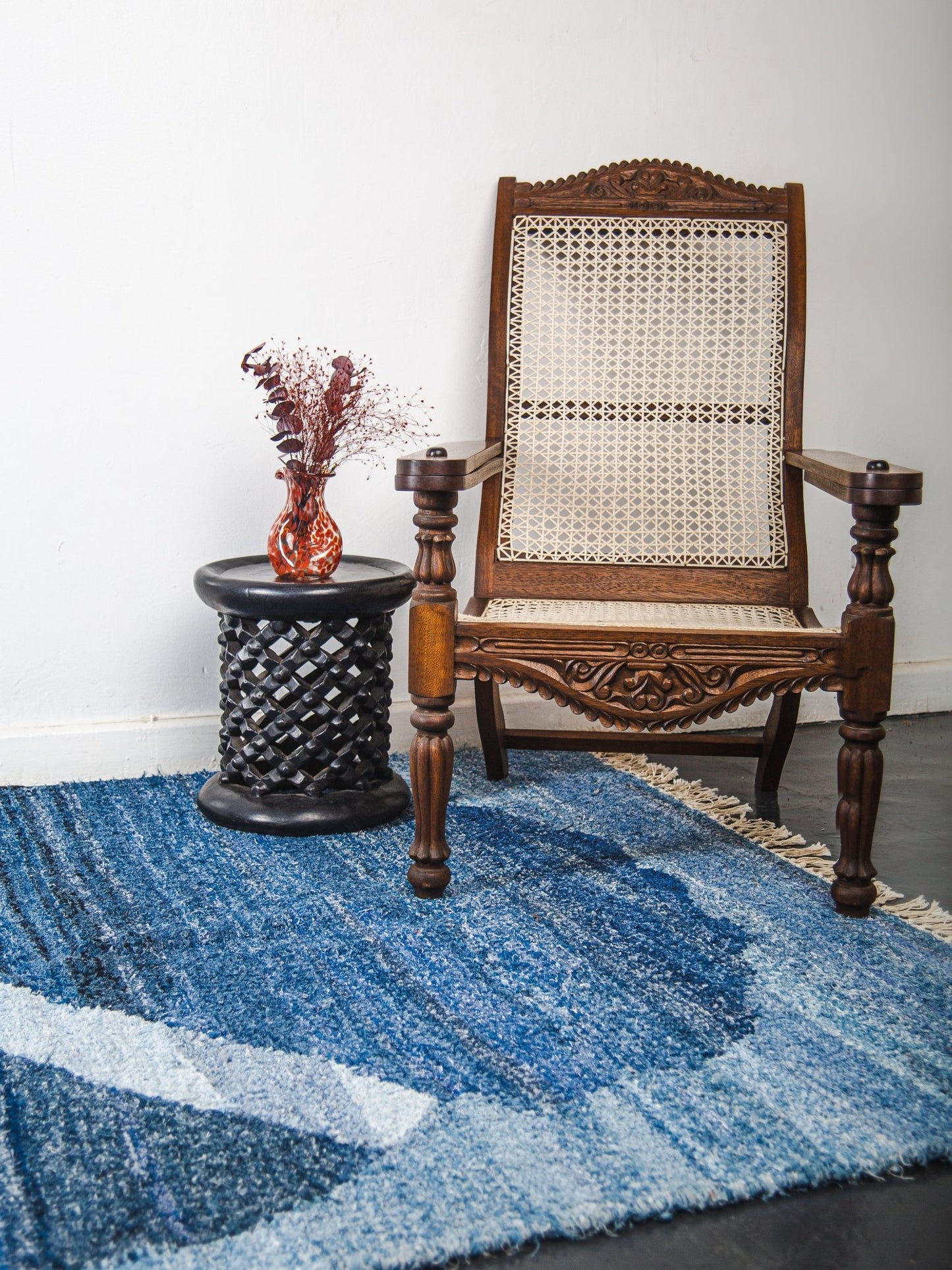 Wooden armchair with woven backrest on a blue patterned rug, next to a black side table with a vase.