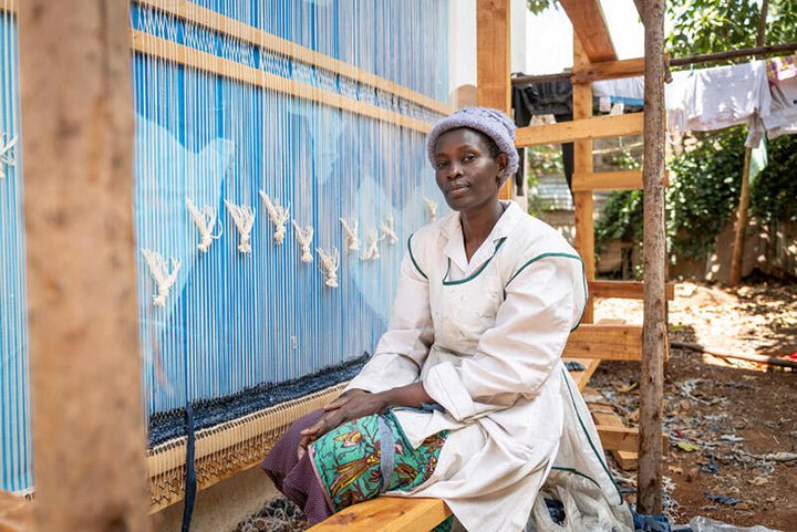Woman sitting in front of a loom with blue fabric, wearing a white shirt and colorful skirt.