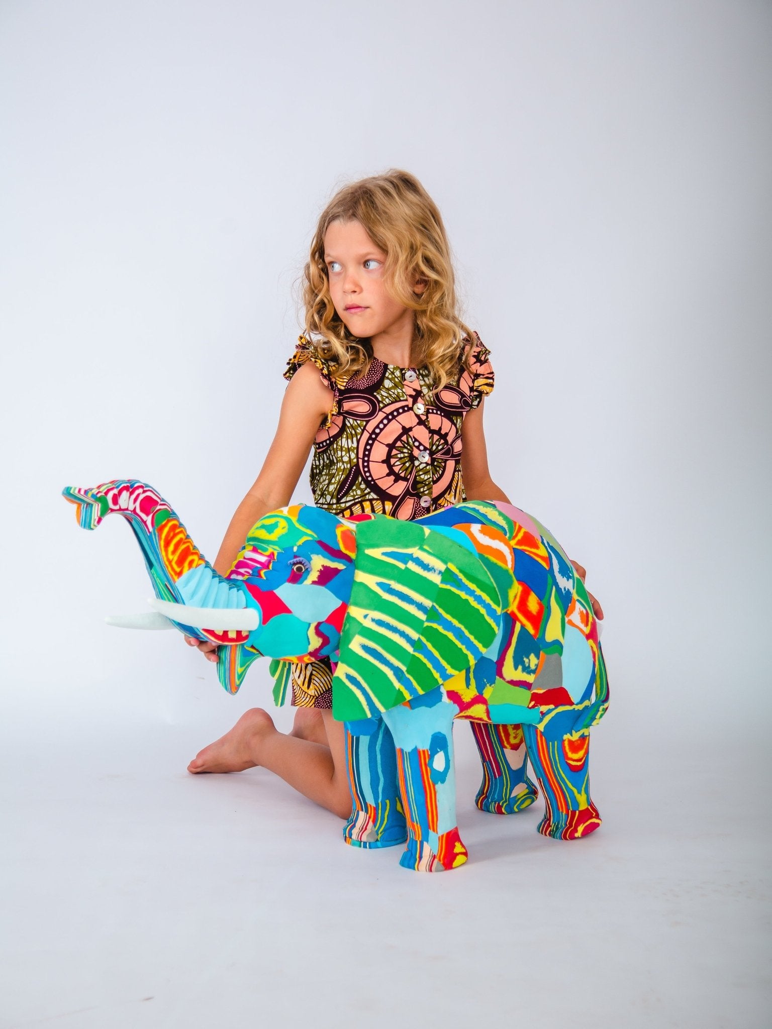 Child sitting on a colourful elephant sculpture against a white background