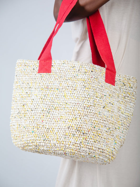 Woven tote bag with red handles held by a person against a neutral background