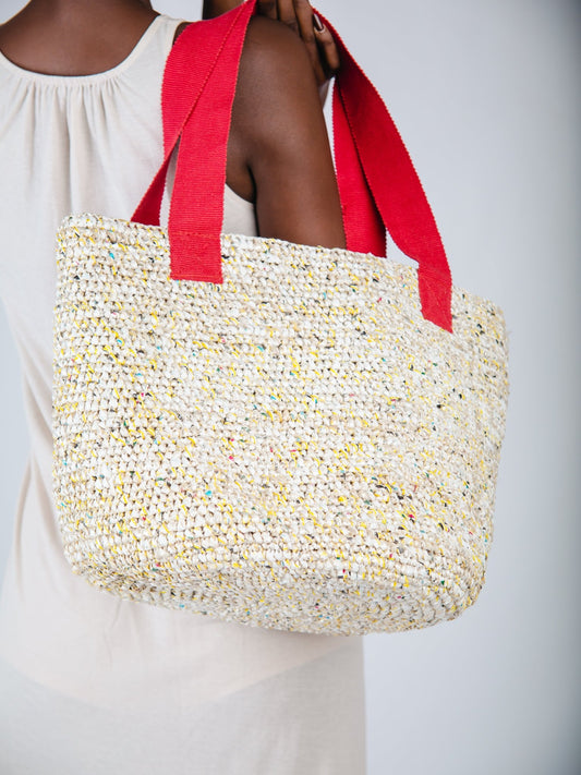 Woven tote bag with red handles held by a person wearing a white dress.