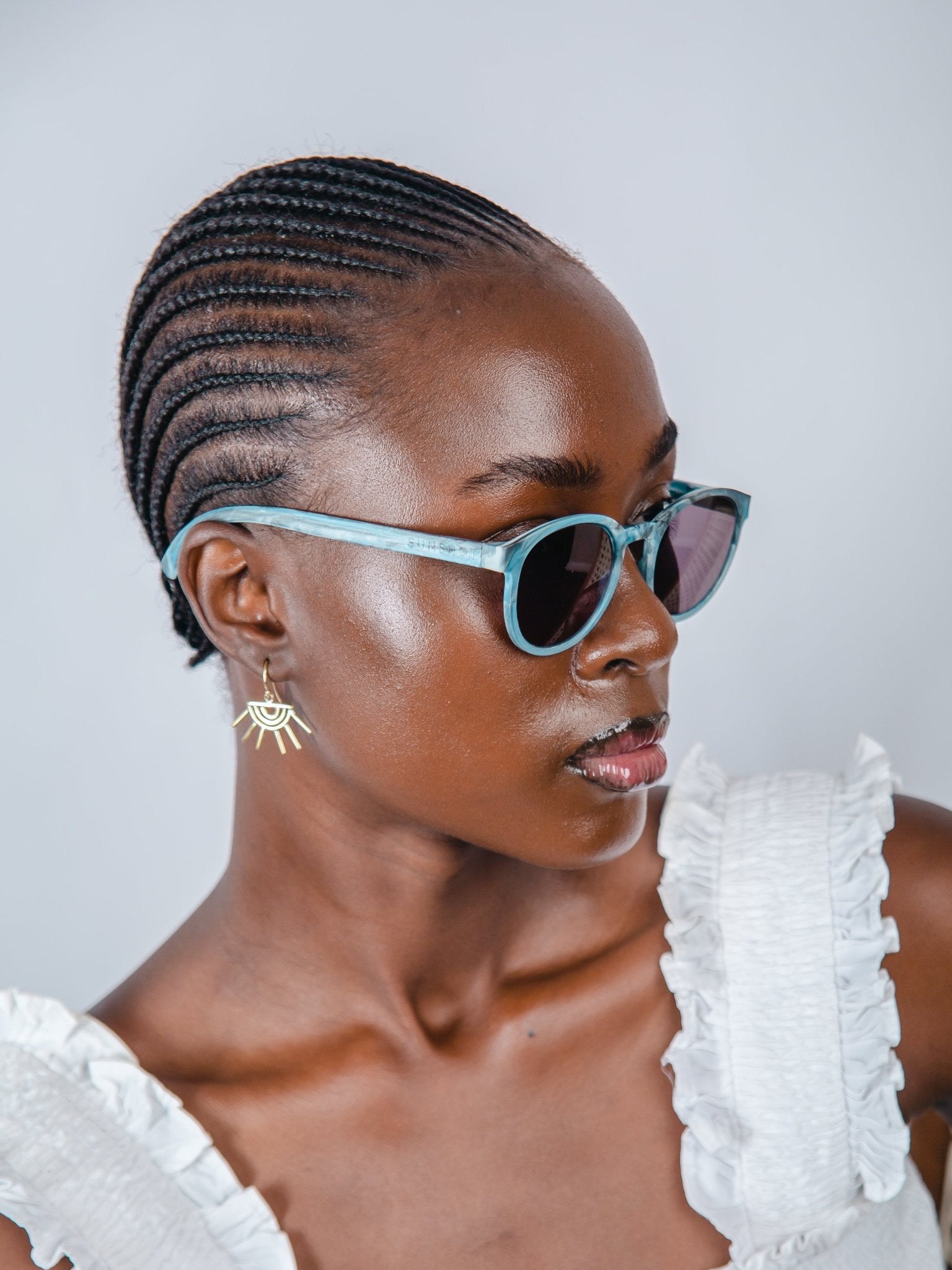 Woman wearing sunglasses and a white top with braided hair against a plain background