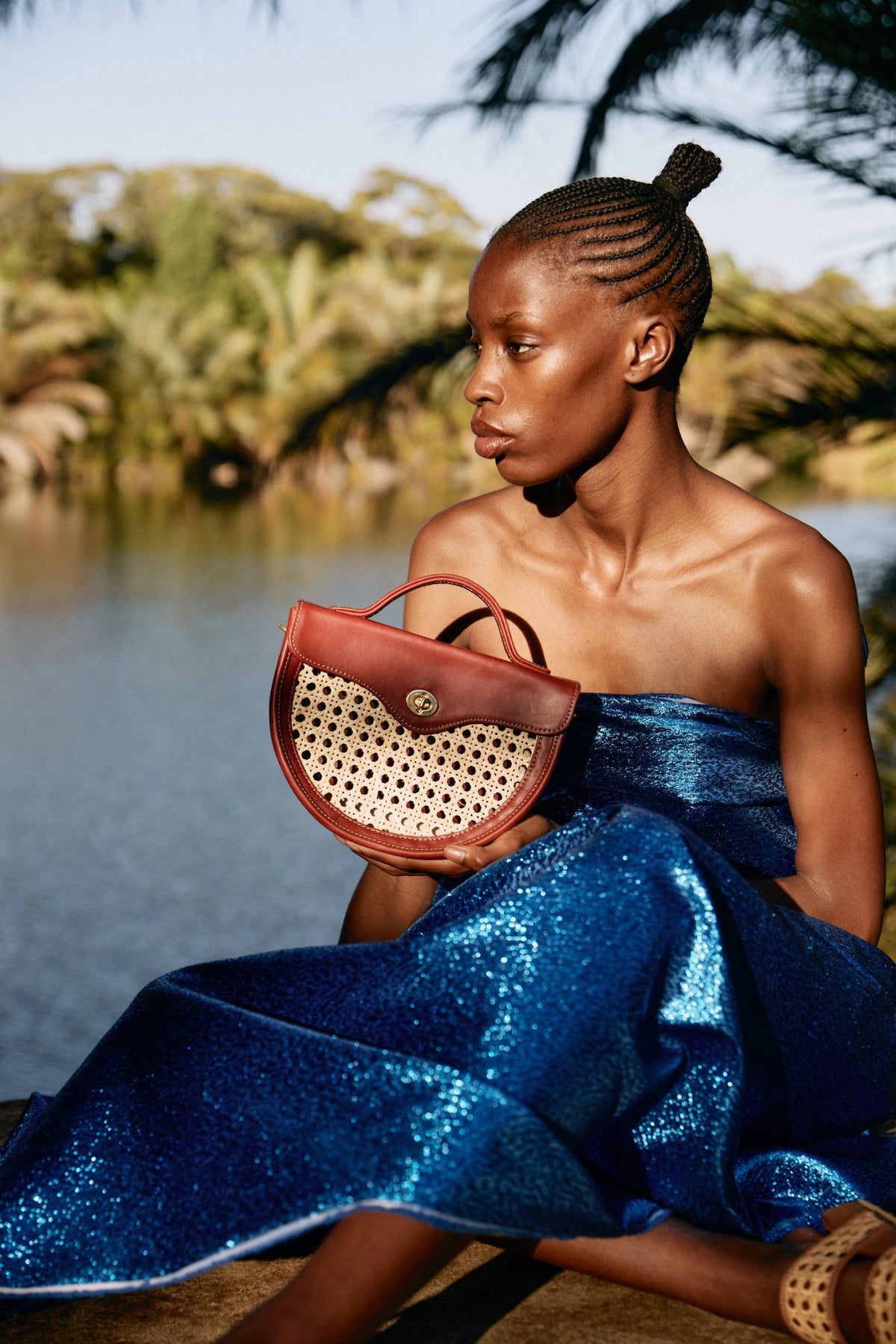 Woman in a blue dress holding a red handbag by a body of water with greenery.
