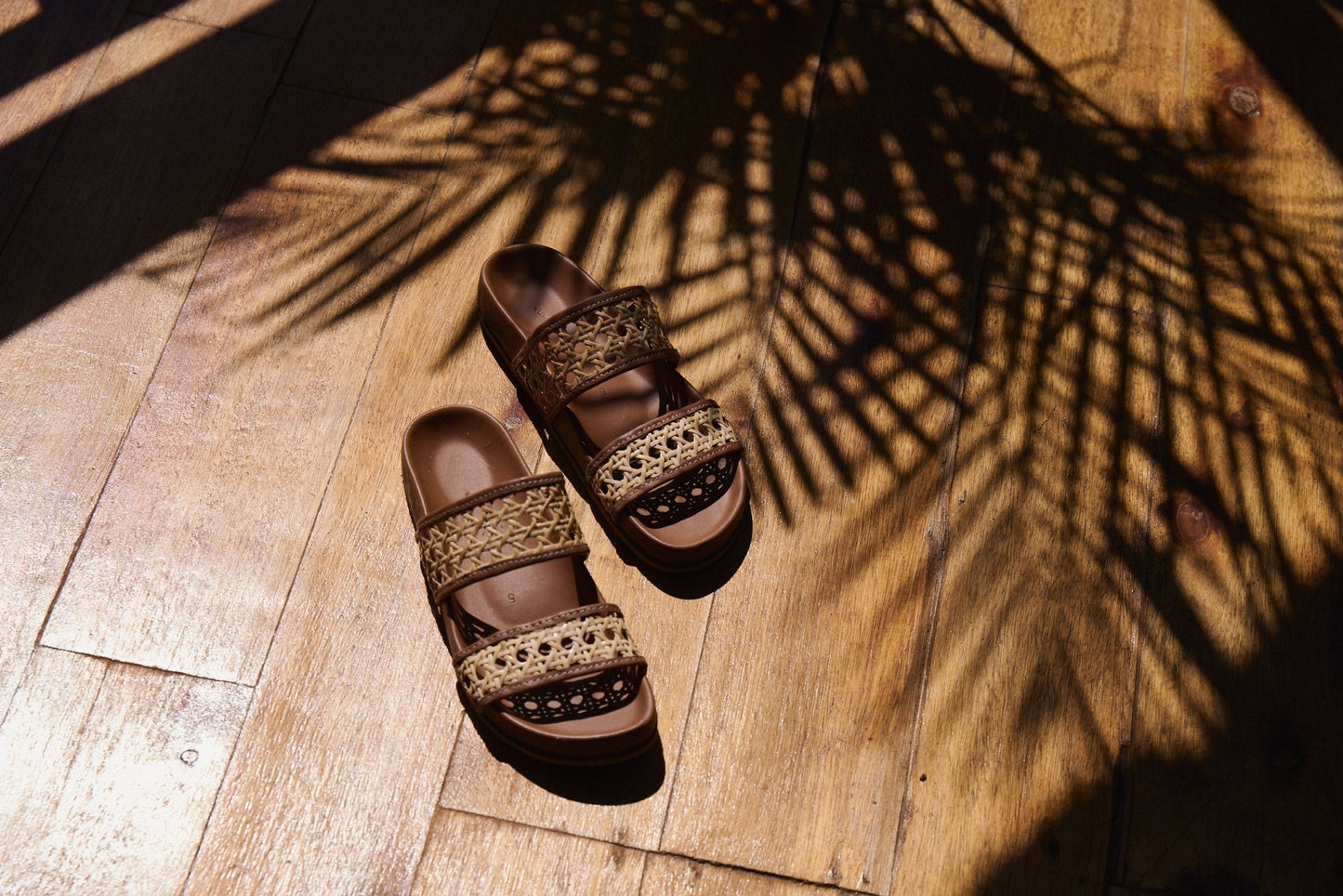 Pair of brown sandals with woven straps on a wooden floor with palm leaf shadows.