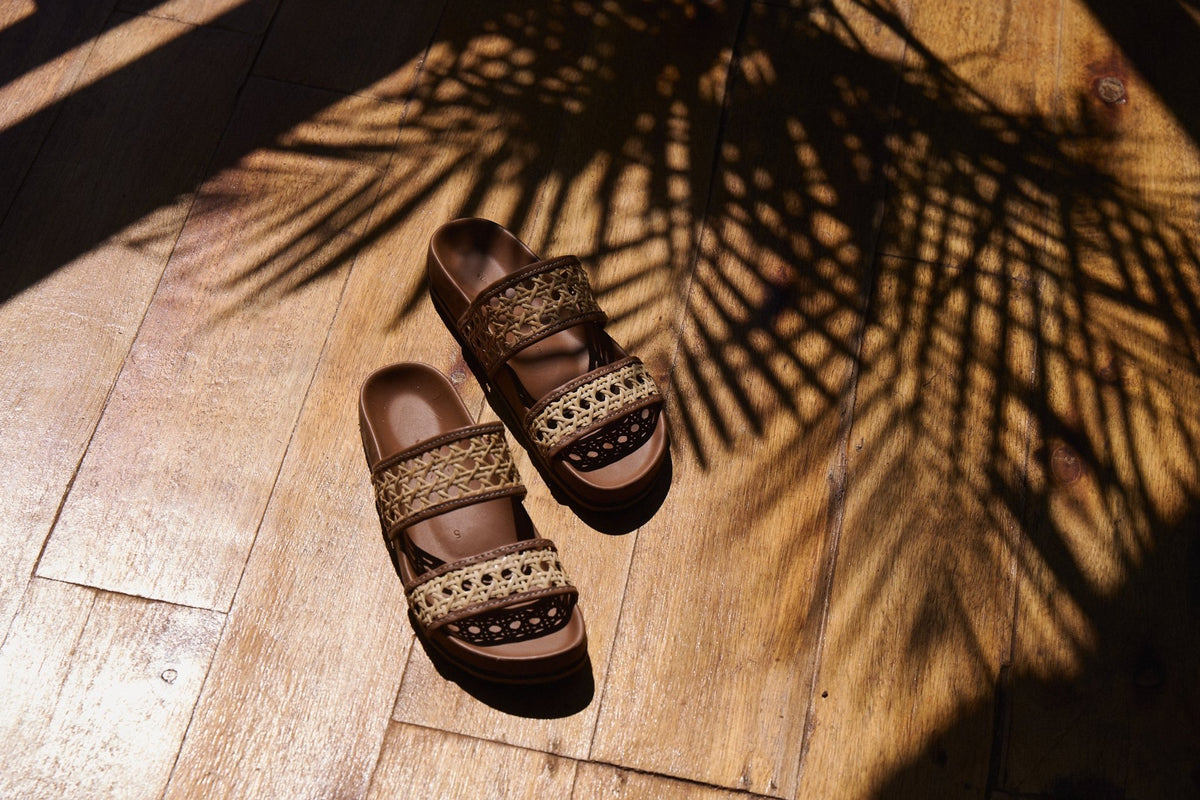 Pair of brown sandals with woven straps on a wooden floor with palm leaf shadows.