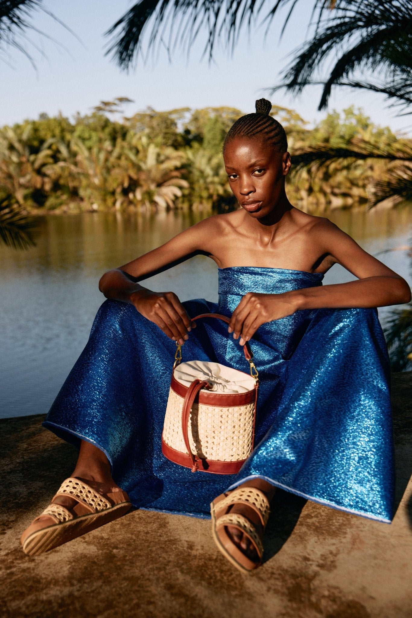 Woman in a blue dress holding a woven bag by a body of water with trees in the background
