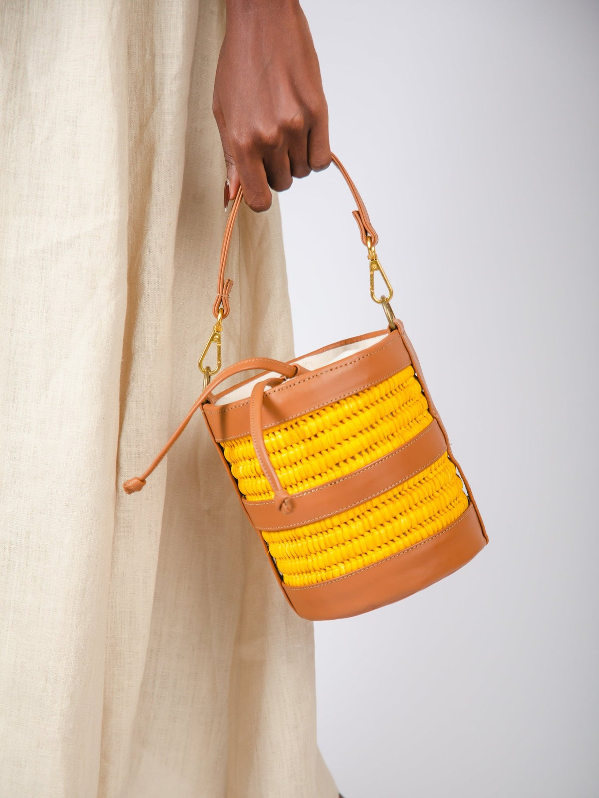 Handbag with yellow woven top and brown leather sides held by a person wearing a beige dress.