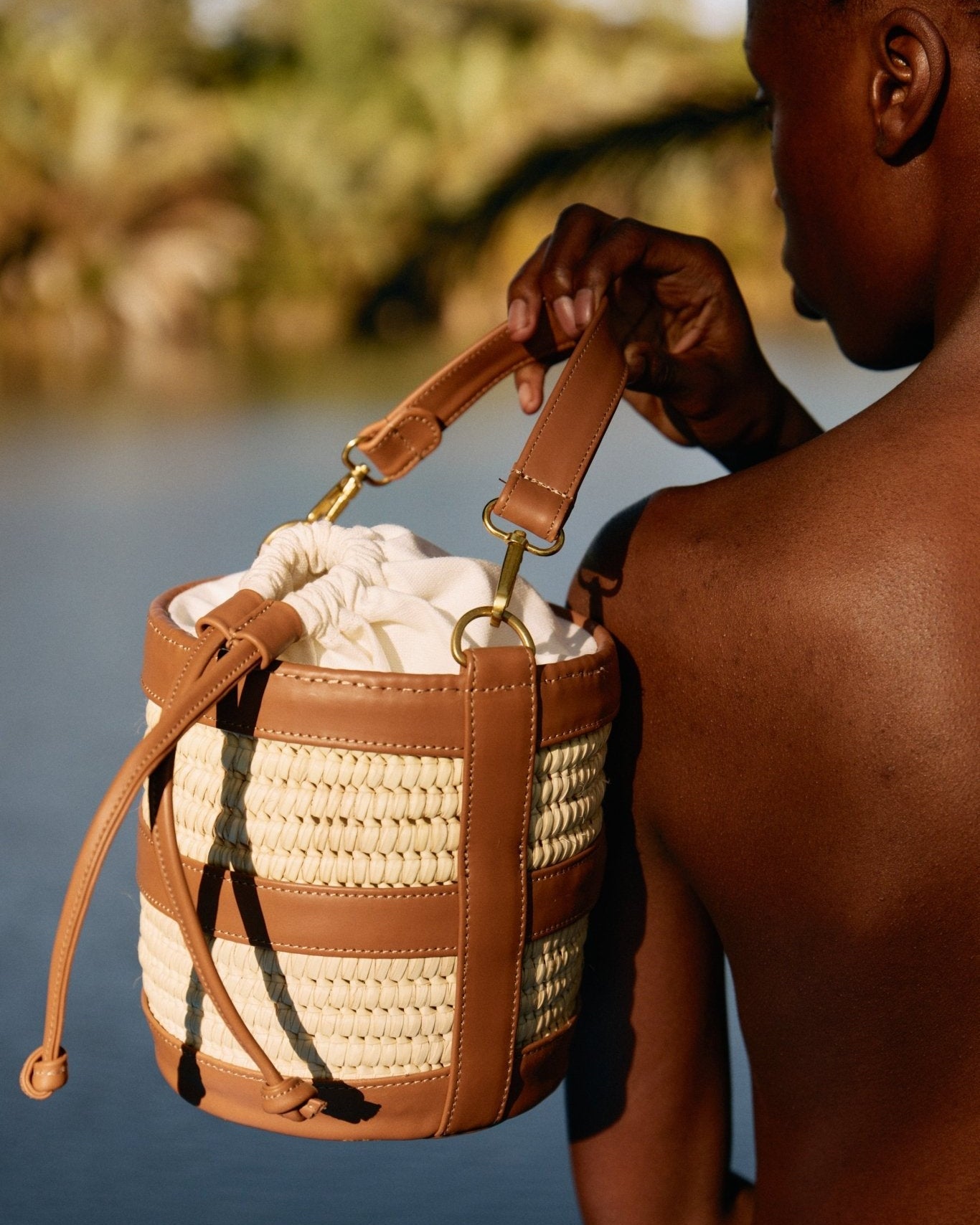 Person holding a woven bucket bag with leather straps by a body of water.