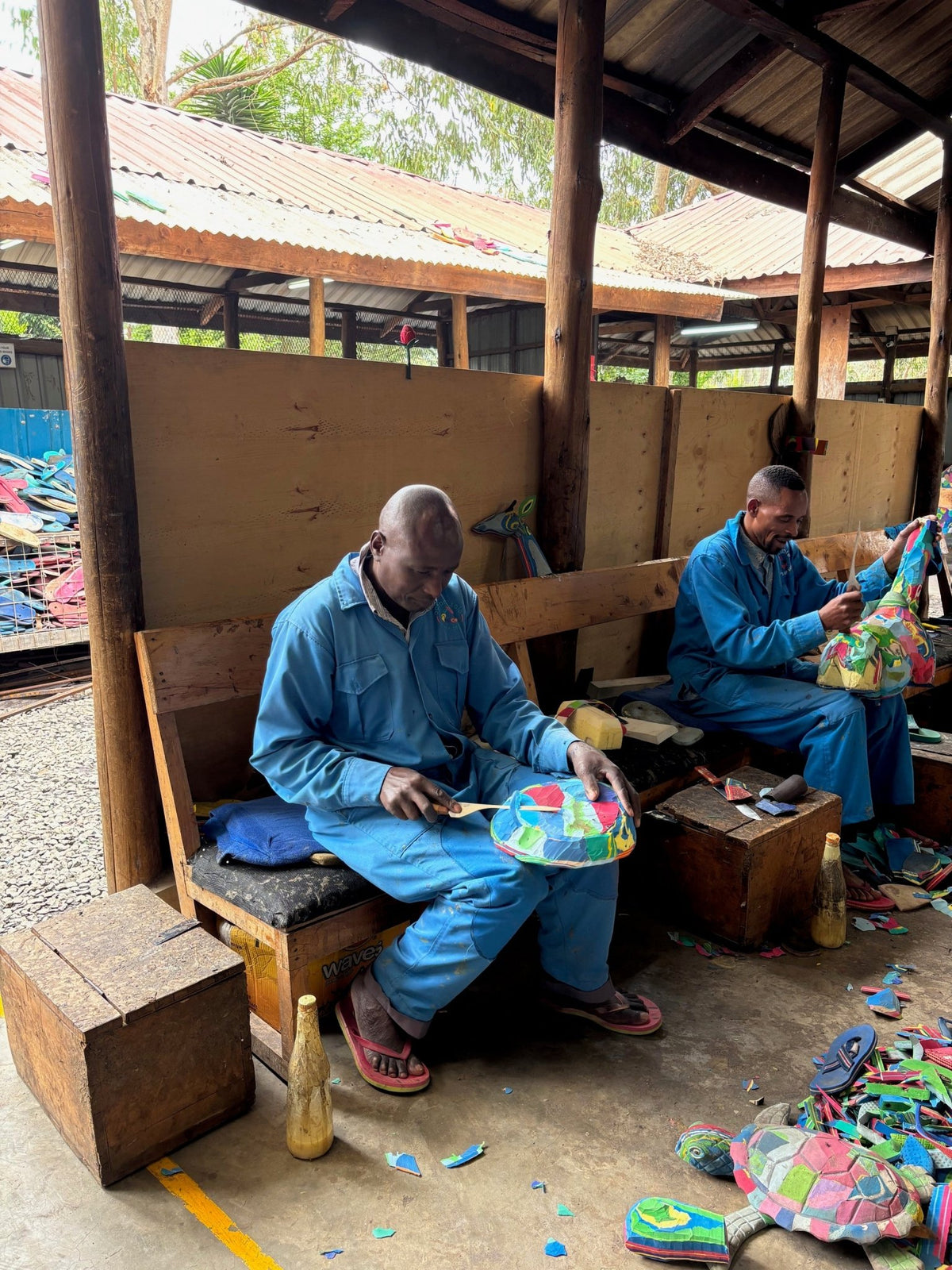 Two individuals in blue uniforms sitting on benches, working with colorful materials in a rustic setting.